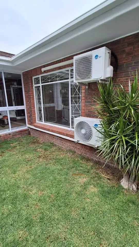 Two aircon outdoor units mounted on a red brick wall in a garden